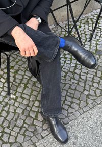 Zwarte leren schoenen met een puntige neus, gedragen met donkerblauwe jeans en een blauwe sok, zittend op een kasseienoppervlak, handen rustend op de knie.
