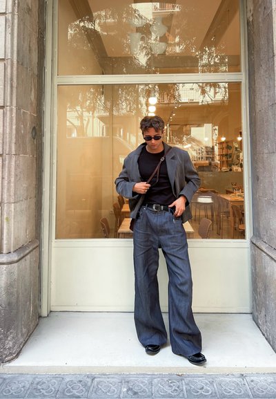 Blazer gris sobre una camisa negra, combinado con pantalones vaqueros de pierna ancha. Los zapatos son negros brillantes, y unas gafas de sol completan el atuendo.