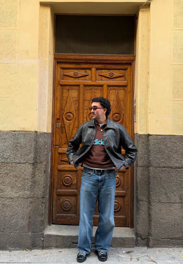 Uomo con occhiali da sole, giacca di pelle nera, felpa Adidas e jeans, in piedi con le mani nelle tasche davanti a una porta in legno ornata.