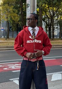 Rdež kapucar na zadrgo z logotipom "scuffers", složen čez belo majico in kravato. Temno jeans hlače, karirasta pas, več prstanov in dodatkov.