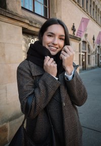 Νεαρή γυναίκα με καρό παλτό προσαρμόζει μαύρο κασκόλ, χαμογελώντας, στέκεται σε πεζοδρόμιο της πόλης με ιστορικά πέτρινα κτίρια στο βάθος.