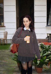 Brun sweater over en hvid skjorte med krave, parret med en plisseret nederdel. Holder en orange rejsekop, stående tæt på grønne planter.