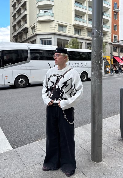 Suéter en blanco y negro con diseño abstracto, pantalones negros holgados, cinturón con detalle de cadena, gafas y gorra, de pie junto a una calle.