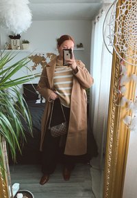 En tanfärgad kappa över en beige randig tröja, svarta byxor och bruna skor. Håller på en rutig väska, med växter och en karta i bakgrunden.