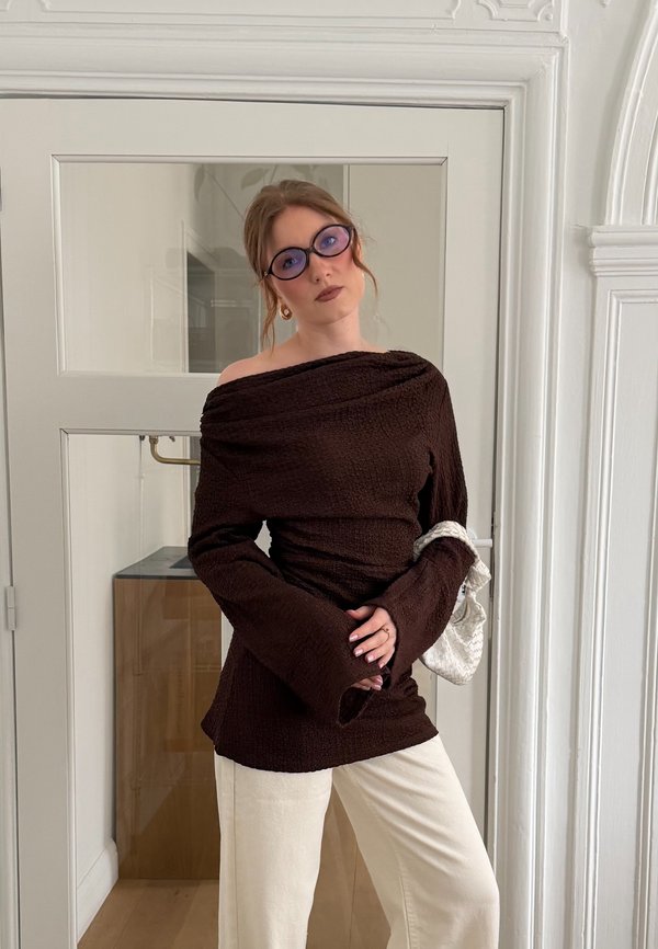 Nainen yllään olkapäät paljastava ruskea neule, kermaiset housut, violettisävyiset silmälasit ja hän pitää kädessään pientä valkoista laukkua valkoisessa sisätilassa.