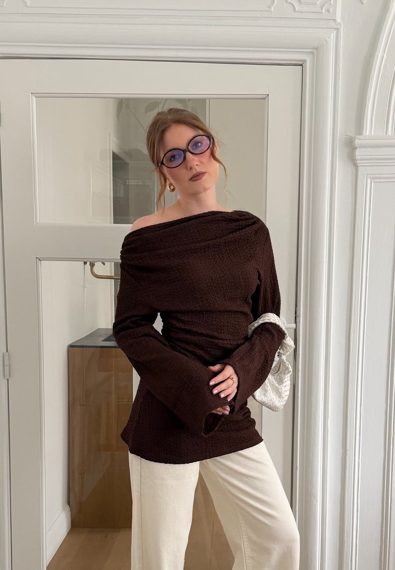 Vrouw die een bruin off-shoulder trui draagt, crèmekleurige broek, paars getinte bril en een kleine witte tas vasthoudt in een witte binnenruimte.