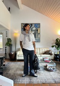 T-shirt blanche, pull gris drapé autour du cou, pantalons à rayures, sac noir et chaussures noires. Intérieur avec un canapé léger et des plantes.