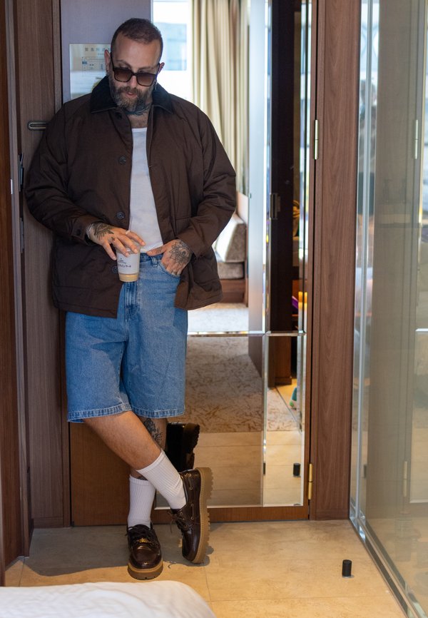 Mann mit Tätowierungen und Sonnenbrille lehnt an der Türöffnung, trägt eine braune Jacke, ein weißes Tanktop, Jeansshorts, weiße Socken und braune Schuhe und hält einen Eiskaffee.