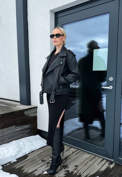 Chaqueta de cuero negra de motorista sobre un suéter de punto negro, combinada con una falda negra ajustada que presenta un corte lateral y botas de tacón negras hasta la rodilla.