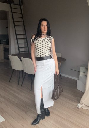 Falda corta blanca combinada con una blusa de lunares. Cinturón negro y botas de vaquero. Bolso marrón. Suelo de madera clara y muebles modernos en el fondo.