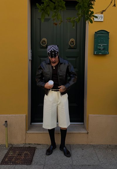 Hombre con chaqueta de cuero negra, pantalones cortos blancos, bandana negra y calcetines sostiene una taza, de pie frente a una puerta verde oscura con un buzón verde.