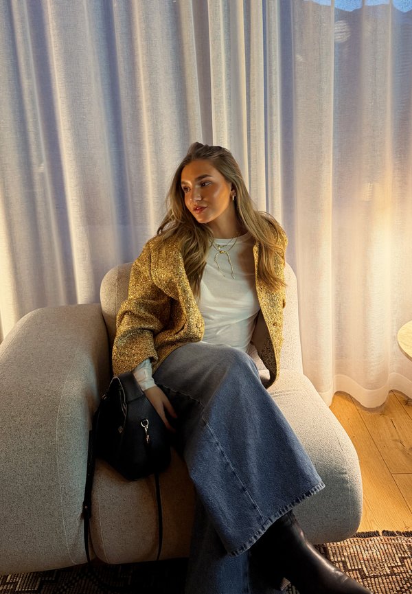 Jeune femme aux longs cheveux portant une veste jaune et une jupe en jean, assise sur un fauteuil beige à côté d'un rideau, tenant un sac à main noir.