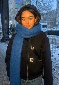 Jeune femme portant une veste noire à capuche et une épaisse écharpe bleue, debout sur un trottoir urbain enneigé avec des voitures et des bâtiments en arrière-plan.