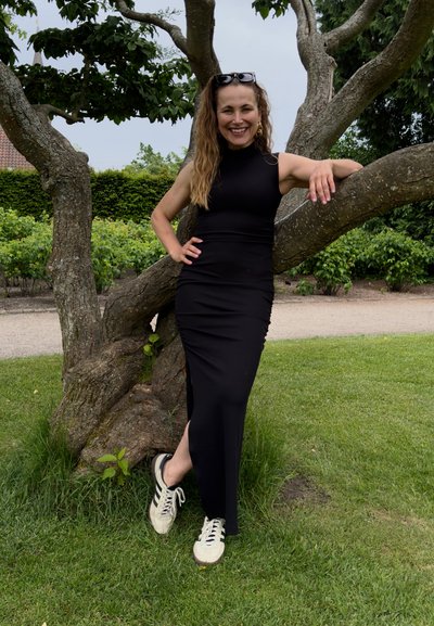 Vestido negro, ajustado, sin mangas con diseño de abertura lateral, combinado con zapatillas blancas. La modelo se apoya contra un árbol en un entorno de parque verde.