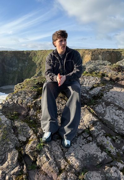 Joven con chaqueta oscura y pantalones grises sentado en un acantilado rocoso cerca del océano bajo un cielo azul con nubes.