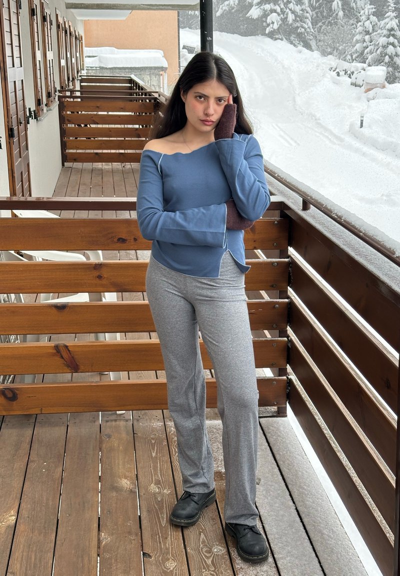 Giovane donna in top blu scivolato sulle spalle e pantaloni grigi in piedi su un balcone di legno accanto a un paesaggio coperto di neve.