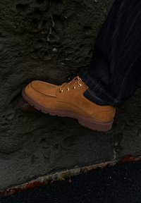 Ghete de gleznă din piele întoarsă maro, cu ochiuri aurii și accente din piele neagră, având o talpă cauciucată texturată. Purtați pe o suprafață stâncoasă.