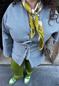 Grey, textured cardigan with a tie waist, worn over green trousers. A patterned yellow scarf and a small leopard-print bag provide additional detail. Green trainers complete the outfit.