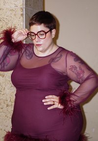 Lühikeste juuste ja prillidega inimene kannab burgundia värvi läbipaistvat kleiti sulgedega varrukate juures ja käel on kassi tätoveering, seisab beežse seinaga vastu.