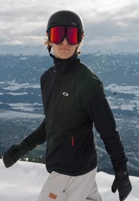 Skiklær med svart glidelåsjakke, lyse bukser, svarte hansker og svart hjelm med røde speilbriller; snødekt fjellbakgrunn.