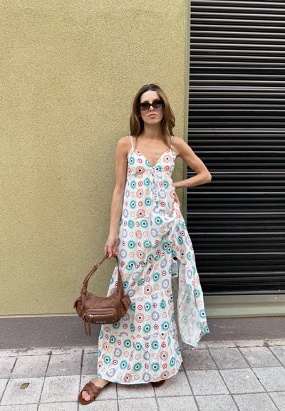 Vestido largo floral con lazos, con un fondo blanco y patrones circulares coloridos en azul, rojo y verde. Bolso marrón y sandalias.
