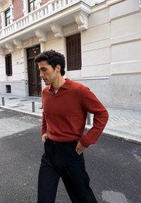 Pull en laine orange rouille avec col en V et manches longues, associé à un pantalon noir. La personne marche sur une rue pavée à côté d'un bâtiment.