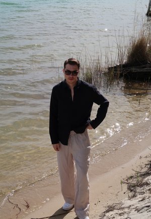 Una persona está de pie en la playa, vistiendo una camisa de botones negra, pantalones beige claros y zapatillas blancas, con gafas de sol y cabello corto.