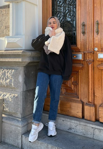 Sudadera negra de gran tamaño, jeans azules y zapatillas blancas. Envuelt@ en una bufanda de piel sintética color crema, de pie junto a una puerta de madera con un diseño intrincado.
