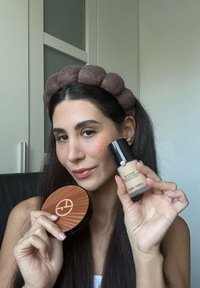 Femme avec de longs cheveux bruns et un bandeau marron tient un flacon de fond de teint Giorgio Armani et un compact dans un environnement intérieur neutre.