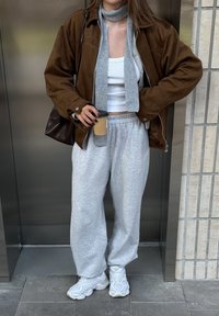Bruine suède jas over een witte crop top, gecombineerd met grijze joggingbroek en witte sneakers. Accessoires zijn onder andere een grijze sjaal en een koffiekopje.