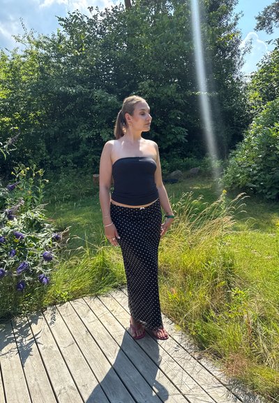 Falda maxi negra con lunares, con un top ajustado negro sin tirantes, sandalias y accesorios; de pie en una terraza de madera rodeada de vegetación.