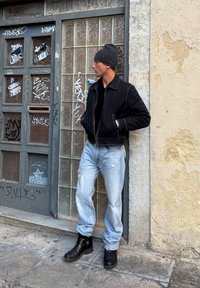 Uomo con giacca nera, berretto, jeans chiari strappati e stivali neri che si appoggia a un muro di vetro coperto di graffiti su un marciapiede urbano.