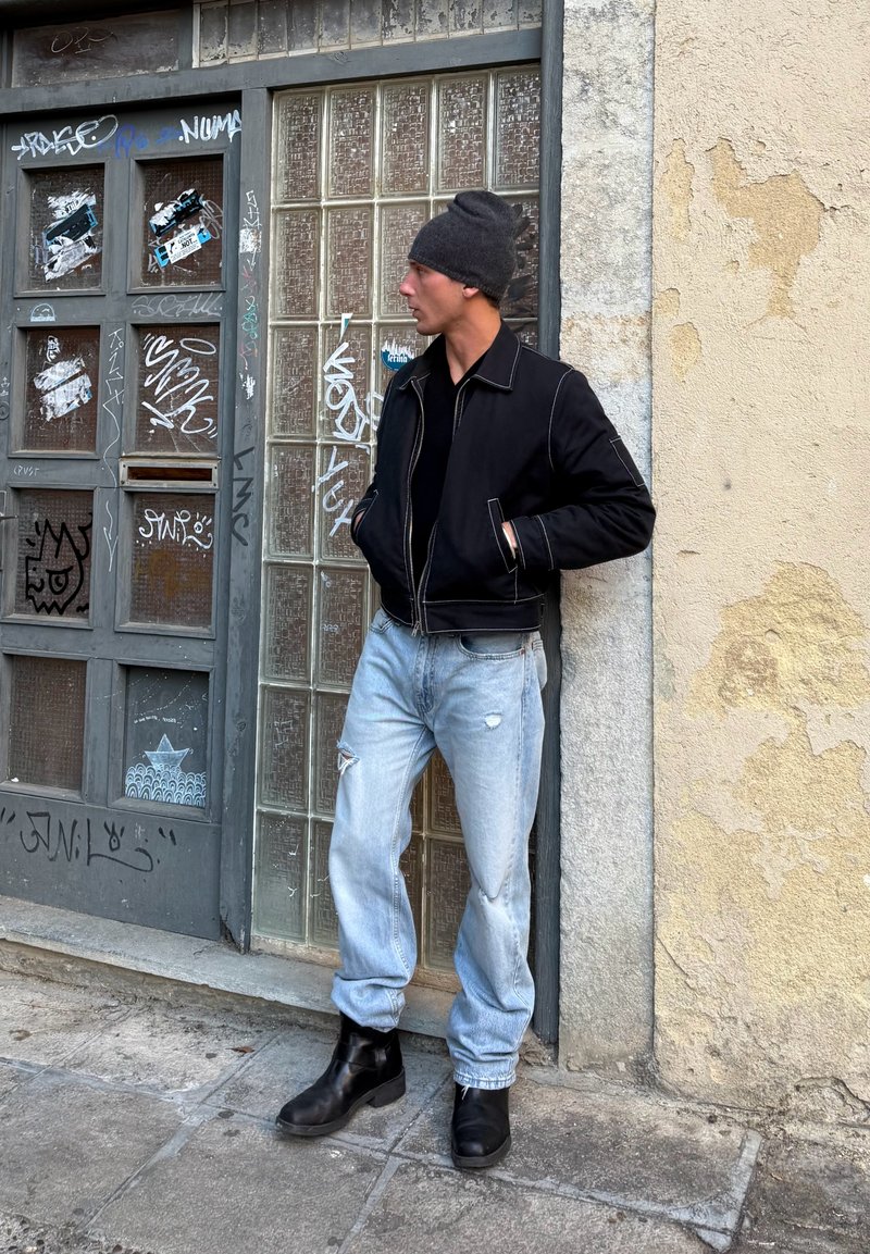 Man som bär svart jacka, mössa, ljusa slitna jeans och svarta stövlar, lutad mot en glasvägg täckt av graffiti på en stadsstående trottoar.