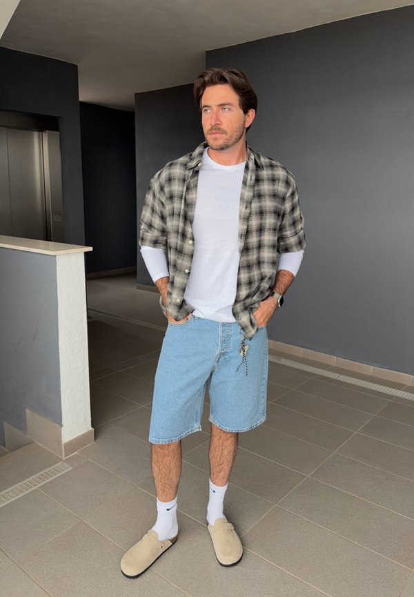 Jeune homme debout à l’intérieur portant une chemise blanche à manches longues sous une chemise à manches courtes à carreaux, un short en denim, des chaussettes blanches et des chaussures beiges sans lacets.