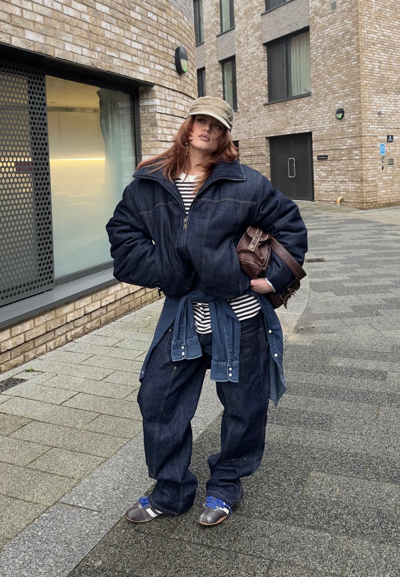 Person mit roten Haaren, die eine übergroße Jeansjacke und Jeans trägt, gestreiftes Hemd, beige Mütze, hält eine braune Tasche, steht auf dem Bürgersteig einer Stadt.