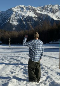 Un homme portant une chemise à carreaux boutonnée et un pantalon vert foncé se tient dans la neige, face à des montagnes enneigées, levant une main pour saluer.
