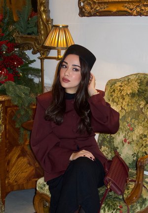 Mujer con abrigo burdeos y sombrero negro sentada en un sillón verde con motivos florales, ajustándose el cabello, con lámpara y decoración festiva de fondo.
