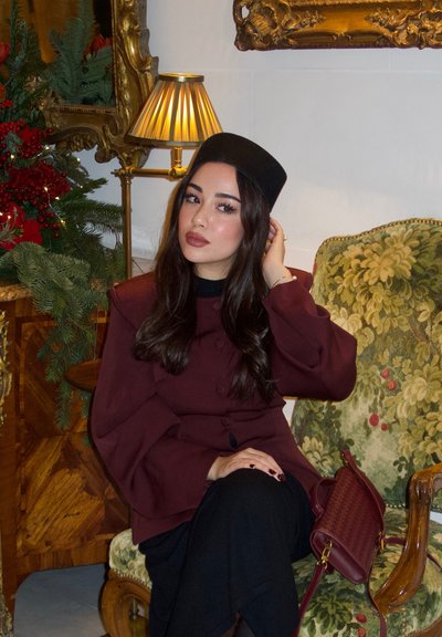 Mujer con abrigo burdeos y sombrero negro sentada en un sillón verde con motivos florales, ajustándose el cabello, con lámpara y decoración festiva de fondo.