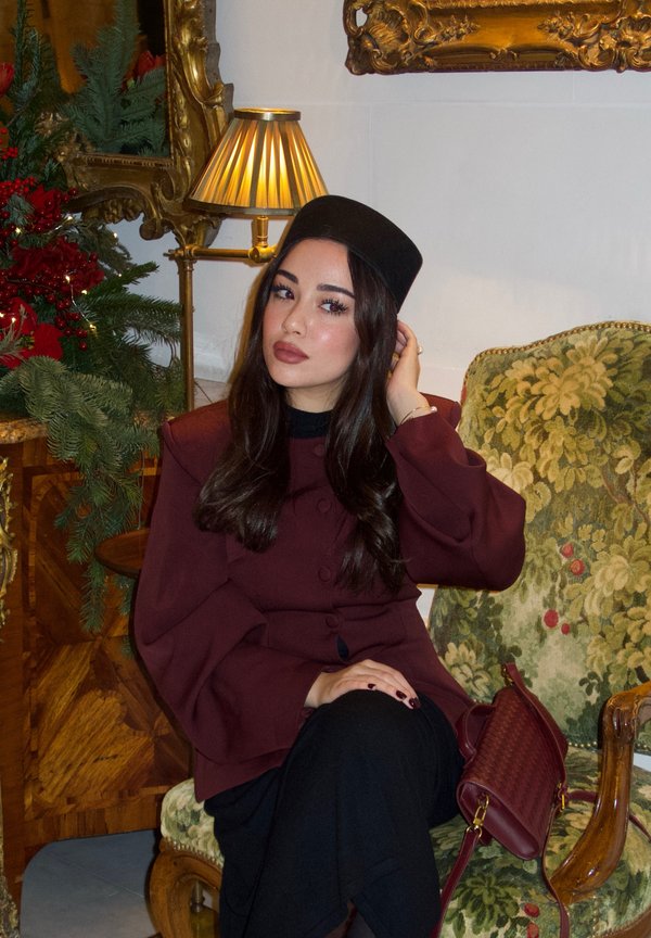 Vrouw in een bordeauxrode jas en zwarte hoed zittend op een groene fauteuil met bloemmotief, haar haar netjes makend, met een lamp en feestelijke groene versiering op de achtergrond.