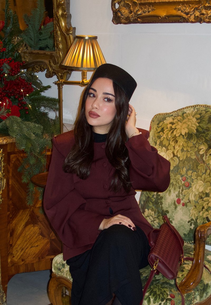 Woman in burgundy coat and black hat sitting on green floral armchair, adjusting hair, with lamp and festive greenery in background.