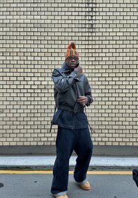 Svart läderjacka med hög krage, oversized mörka jeans och en färgglad randig hatt. Bakgrunden har en tegelvägg.