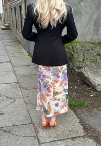 Blazer negro a medida, falda maxi de satén floral con patrones en naranja y púrpura, y zapatos de tacón altos naranjas contra un fondo de acera.