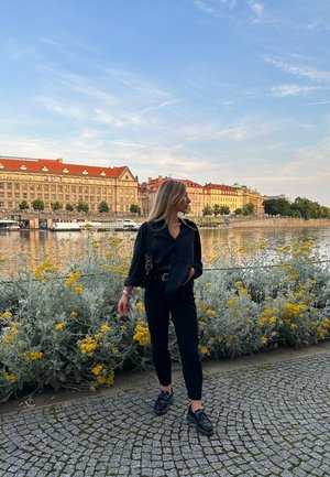 Camisa negra de botones y pantalones combinados con zapatos negros, frente a un río con flores amarillas y edificios históricos de fondo.