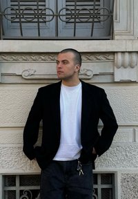 Blazer preto sobre uma t-shirt branca, combinado com jeans escuros. O contraste de texturas com uma parede texturizada e detalhes arquitetónicos ornamentados ao fundo.