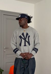 Mann med mørk hud som har på seg en grå genser og mørkeblå caps med NY-logo, står inne med hendene foldet foran en lukket dør.