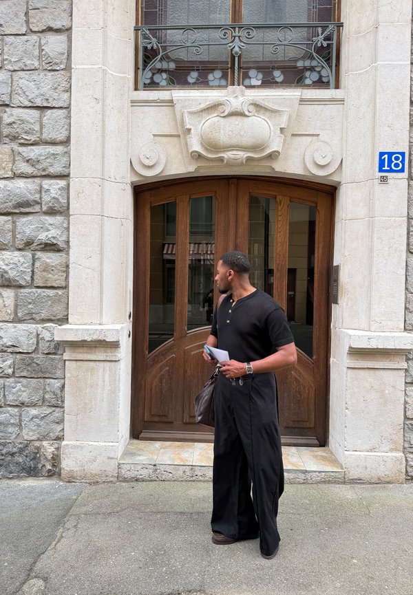 Άνδρας με μαύρη στολή που κρατάει χαρτιά και τσάντα στέκεται μπροστά σε περίτεχνη ξύλινη πόρτα με πέτρινο πλαίσιο και μπλε πινακίδα με τον αριθμό 18.