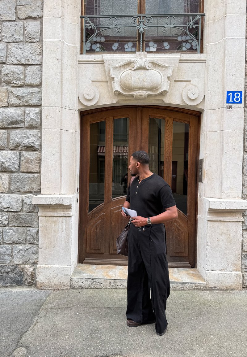 Mężczyzna w czarnym ubraniu trzymający papiery i torbę stoi przed ozdobionymi drewnianymi drzwiami z kamienną ramą i niebieską tabliczką z numerem 18.