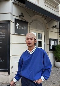 Veste légère bleue à fermeture éclair, avec des poignets et un col à rayures blanches ; portée sur un jean foncé. En arrière-plan, un panneau indiquant "KITCHEN LIBRARY".