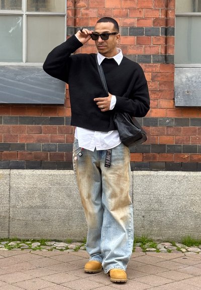 Suéter negro de gran tamaño, camisa blanca con cuello debajo, jeans claros desgastados, zuecos color beige, bolso cruzado negro, gafas de sol.