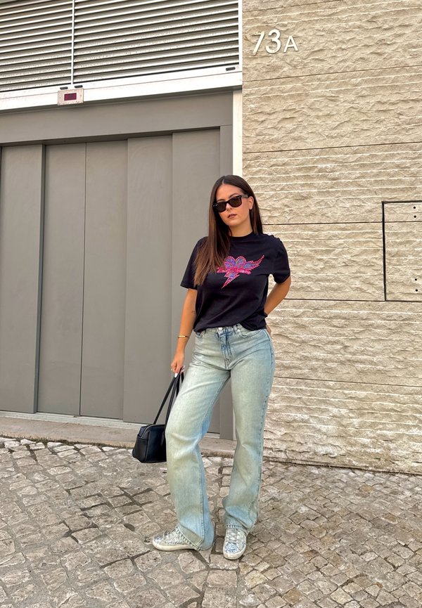 Femme portant des lunettes de soleil, un t-shirt noir à motif graphique, un jean bleu clair, des chaussures argentées, tenant un sac à main noir, se tenant dans une rue pavée près du bâtiment numéro 73A.